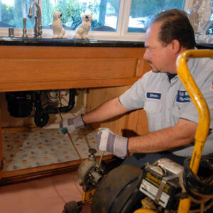 Home - Bill Howe A plumber looks under a kitchen sink to fix a common plumbing problem.